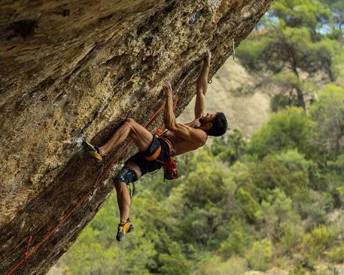 Male rock climber ascending a steep outdoor cliff with greenery in the background.