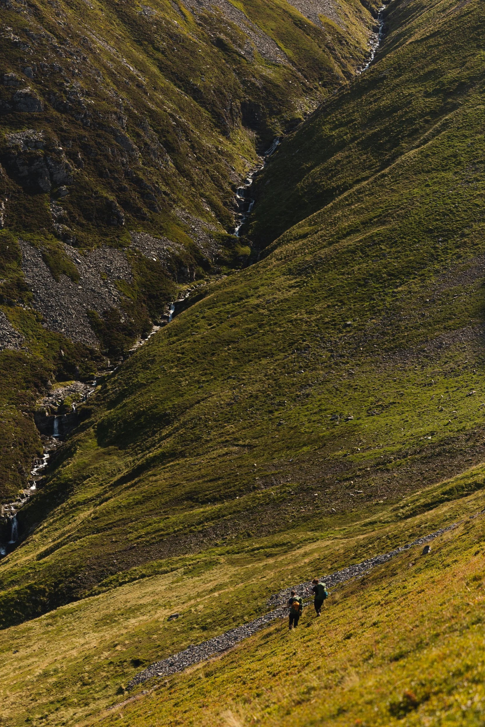 Two hikers walking along a trail in a lush, green mountain valley.