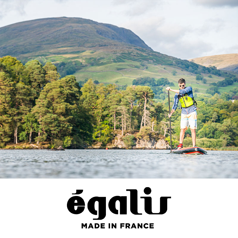 Man stand-up paddleboarding on a lake with lush green trees and mountains in the background.
