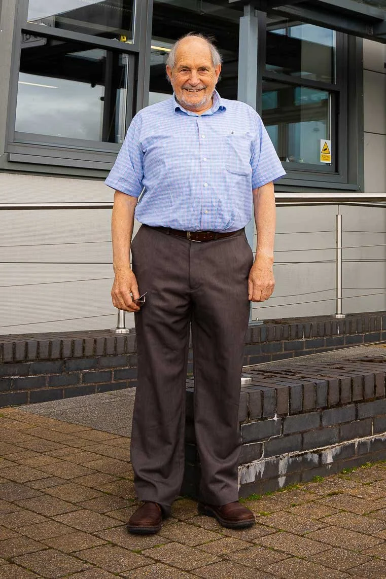An elderly man with gray hair and a beard, wearing a light blue checkered short-sleeve shirt and dark gray pants, standing outdoors on a paved sidewalk in front of a modern building with large glass windows.