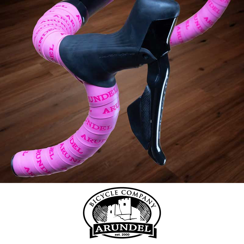 Close-up of a bicycle's handlebar with pink branded tape reading 'ARUNDEL' and a black aero handlebar shifter, against a wooden floor background, with a logo of Bicycle Company Arundel at the bottom.