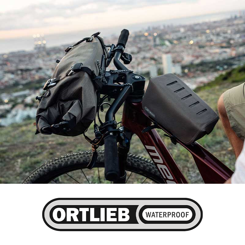 A mountain bike with a black handlebar bag and a black rectangular top tube bag, parked on a hillside overlooking a city during dusk.