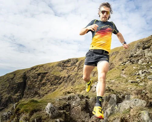 Man running downhill on rocky terrain in outdoor setting, wearing athletic gear and sunglasses.