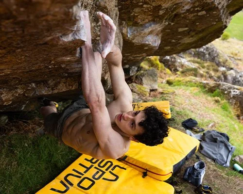 A young man with curly hair rock climbing outdoors, reaching for handholds on a large overhanging rock, with climbing gear and a yellow crash pad on the ground.