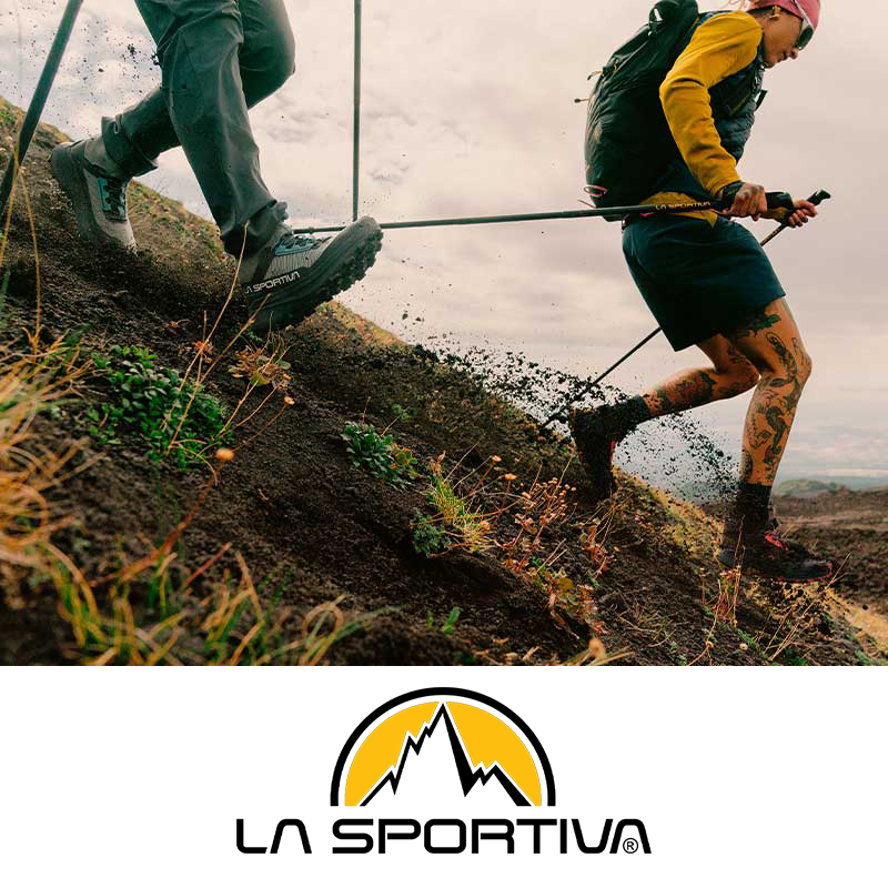 Two people hiking up a steep dirt trail, one wearing gray pants, gray and blue shoes, and the other in a yellow jacket, dark shorts, and tattoos on their legs.