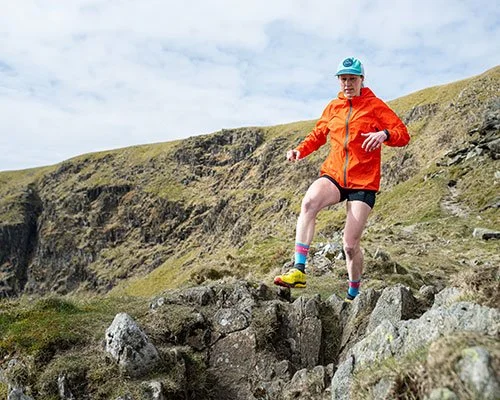 Person in an orange jacket, black shorts, and colorful socks jumping over rocks in a grassy, hilly outdoor landscape.