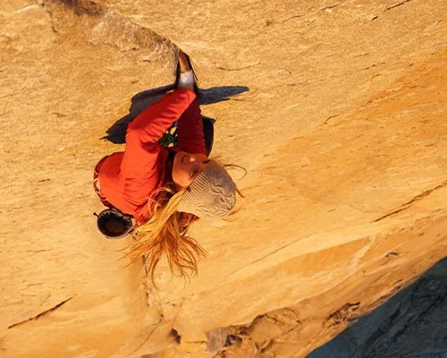 A woman rock climbing on a steep, sandy, orange cliff face with long blonde hair. She is wearing a gray knit hat, red jacket, and black climbing gear.