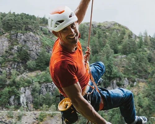 Man wearing a helmet rock climbing outdoors with trees and rocky terrain in the background.