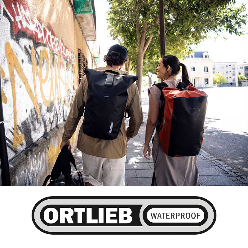 Two people walking on a sidewalk with backpacks, one carrying a bag, near a graffiti-covered wall and trees, in an urban setting.