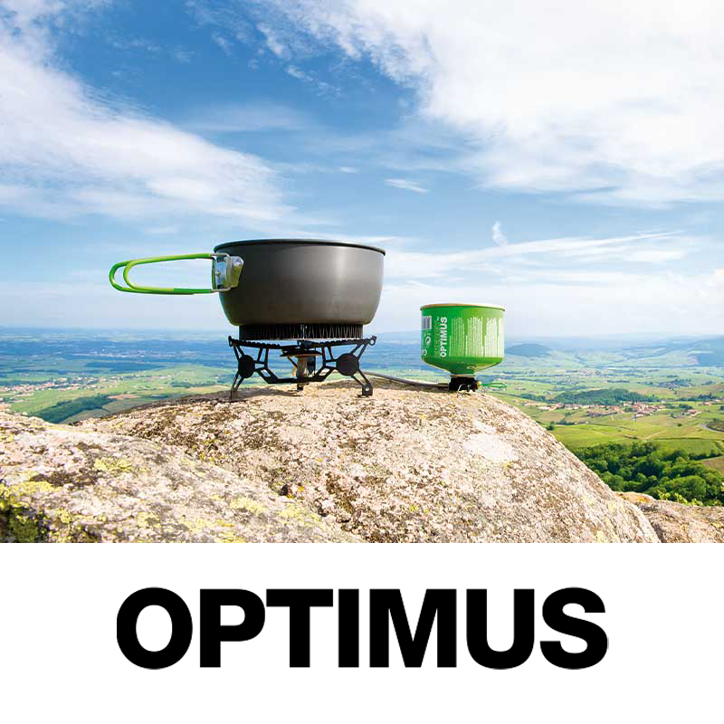 A camping stove with a black pot and a green container on a large rock, overlooking a green landscape and blue sky with clouds.