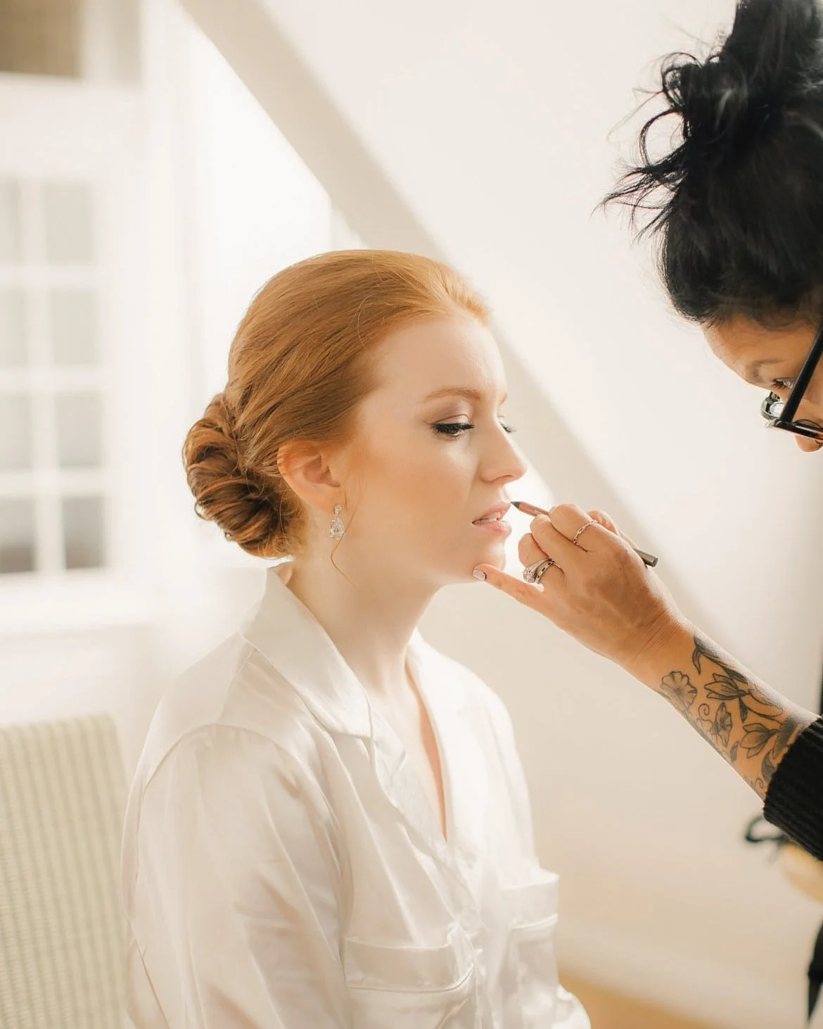 A woman with red hair styled in an elegant updo getting her makeup done by a makeup artist with tattoos on her arm.