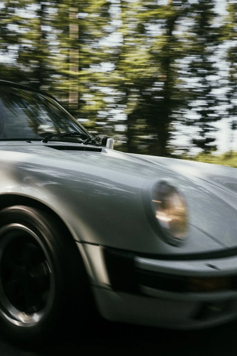 Nahaufnahme eines sportlichen, silbernen Autos in Bewegung auf einer Straße, im Hintergrund sind Bäume und Himmel zu sehen.