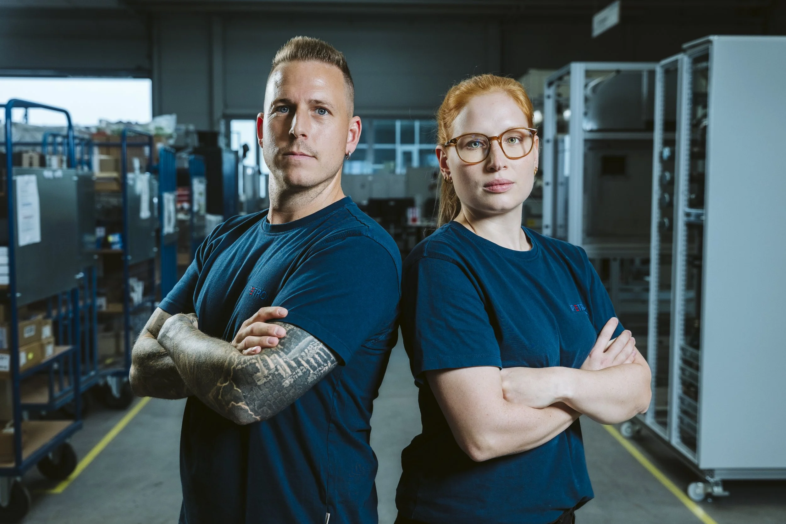 Zwei Personen stehen mit verschränkten Armen in einem Lagerraum, umgeben von Regalen mit elektronischen Geräten und Kisten.