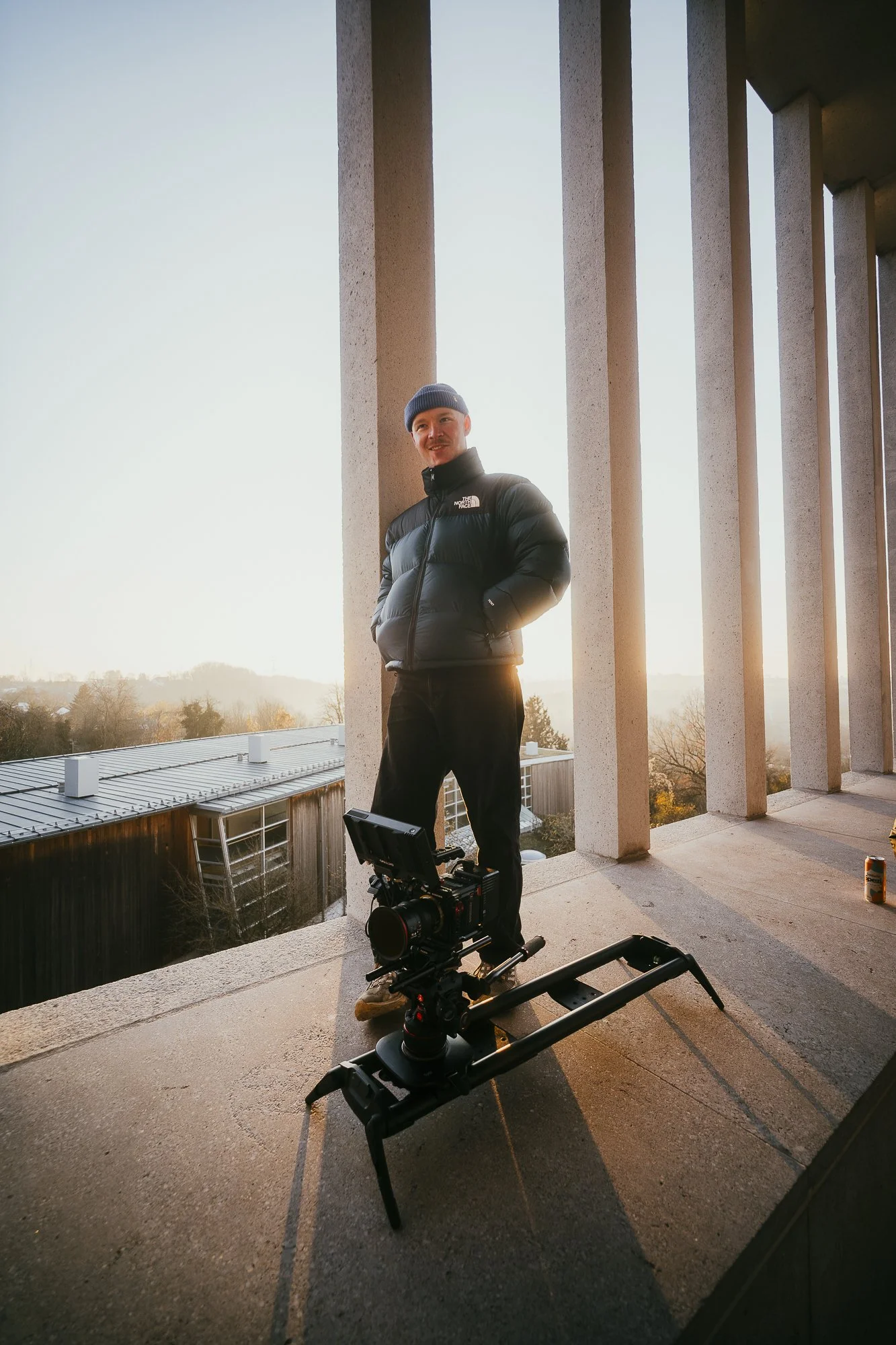 Ein Mann steht auf einer Veranda vor mehreren großen, vertikalen Säulen, mit einem Filmkamera-Setup im Vordergrund, während die Sonne im Hintergrund untergeht.