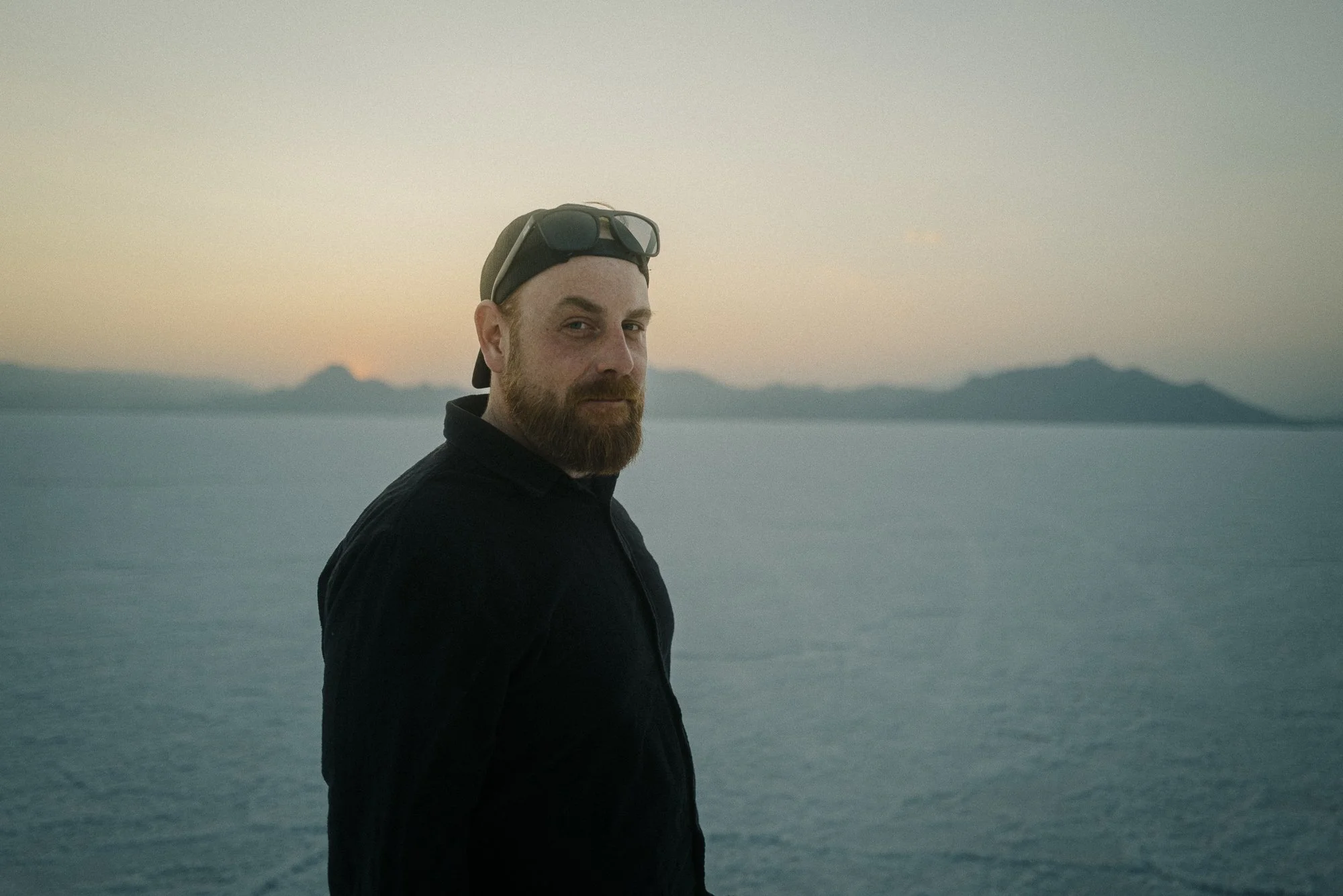 Ein Mann mit Brille auf dem Kopf steht am Wasser bei Sonnenuntergang. Hinter ihm sind Berge sichtbar.