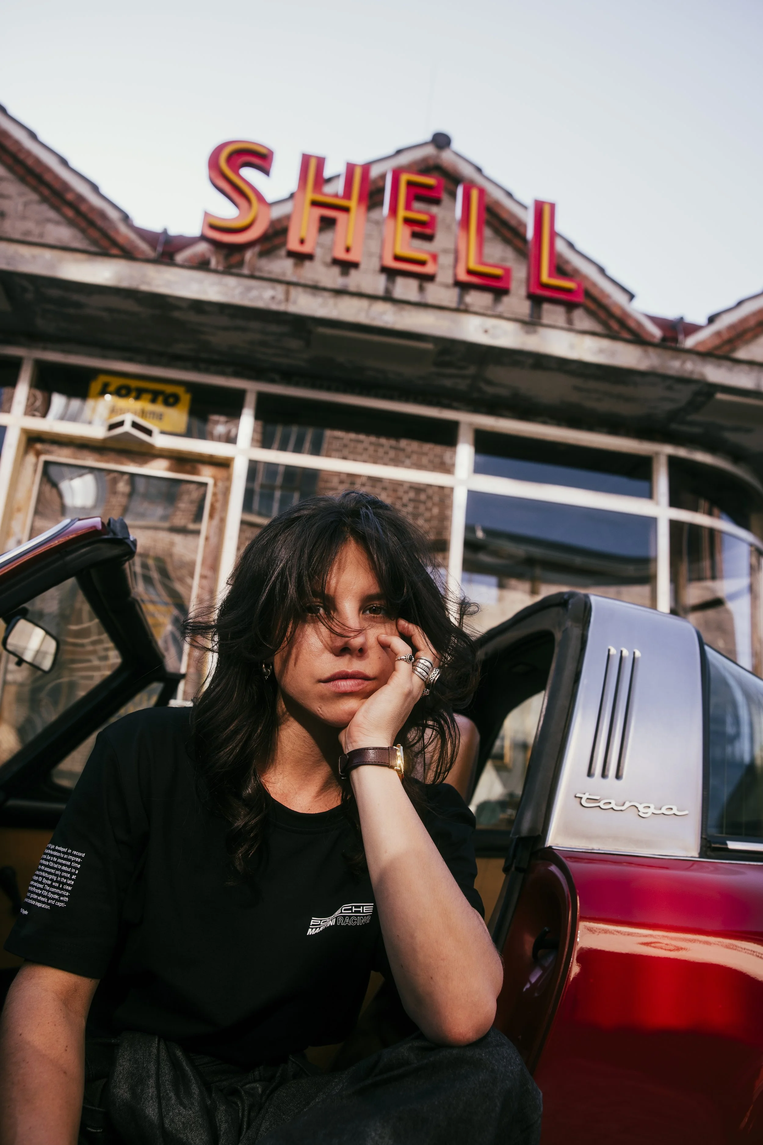 Junge Frau mit dunklen Haaren und schwarzem T-Shirt sitzt neben einem roten Targa Cabrio vor einem Shell-Restaurant.