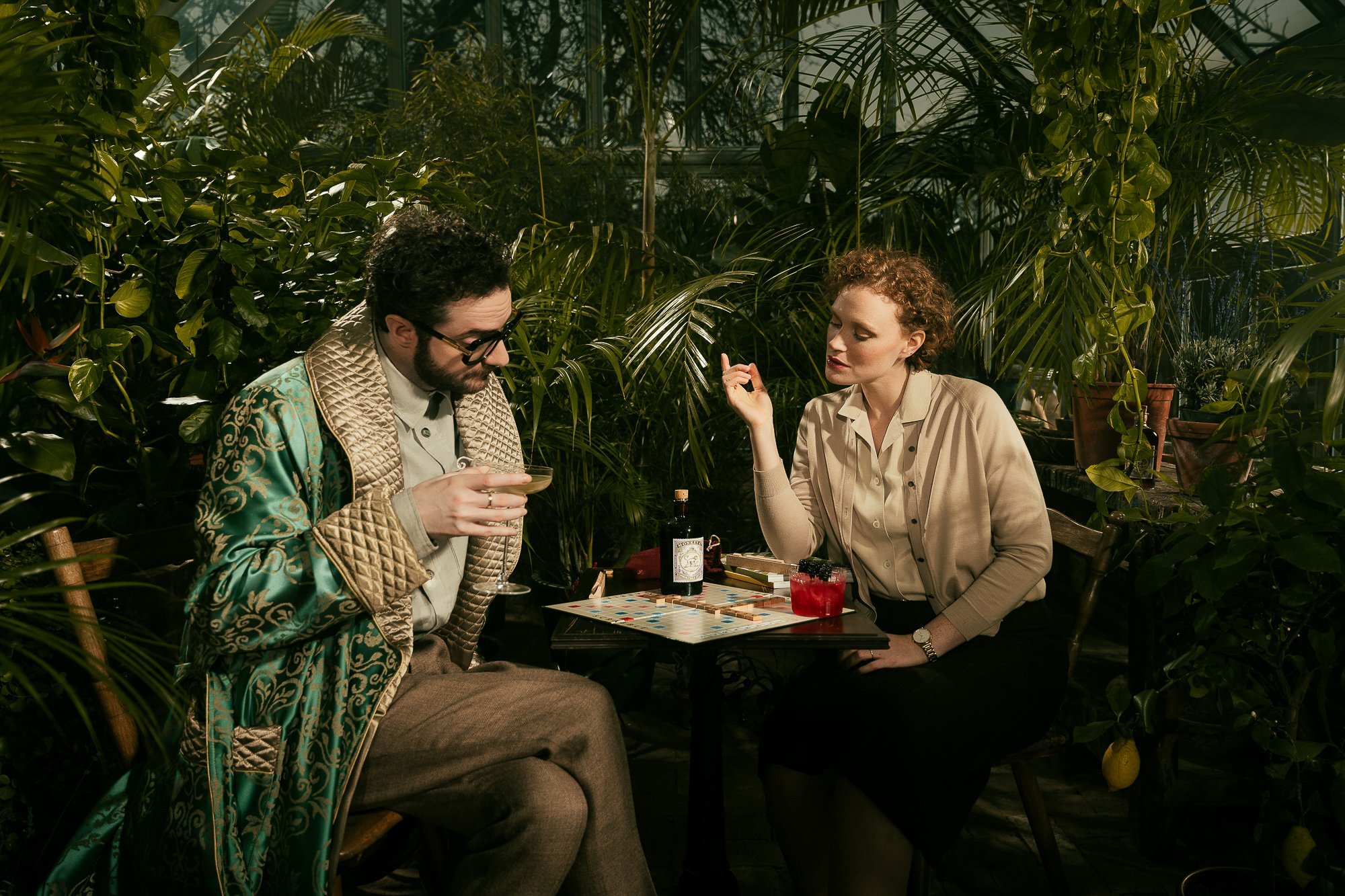 Zwei Menschen sitzen an einem Tisch in einem Gewächshaus umgeben von Pflanzen, sie spielen ein Brettspiel und trinken Getränke.