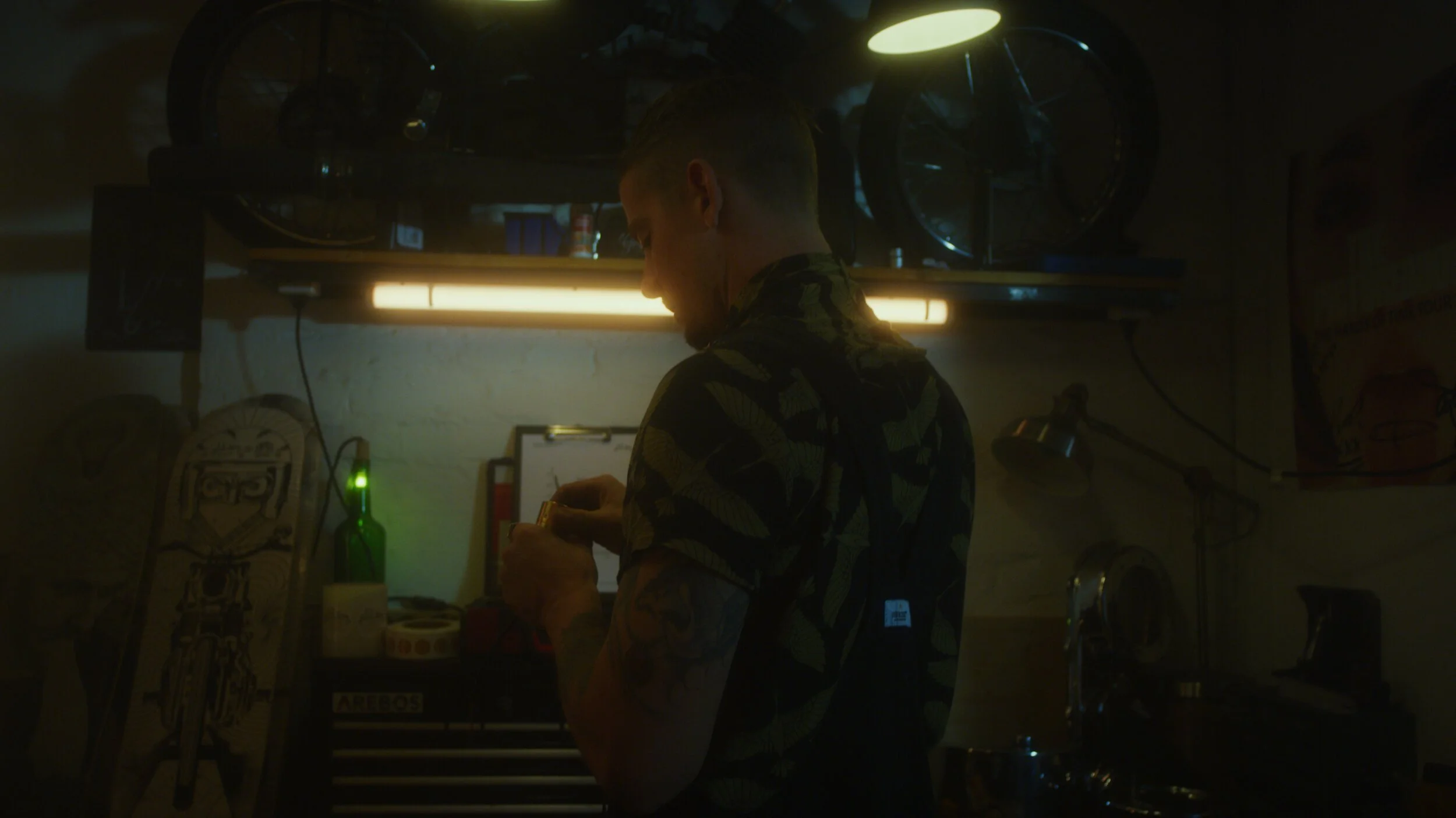 Junge Person in einem Raum mit dunkler Beleuchtung, die an einem Werkstatt- oder Hobbybereich arbeitet, umgeben von Werkzeugen und Ausrüstung.