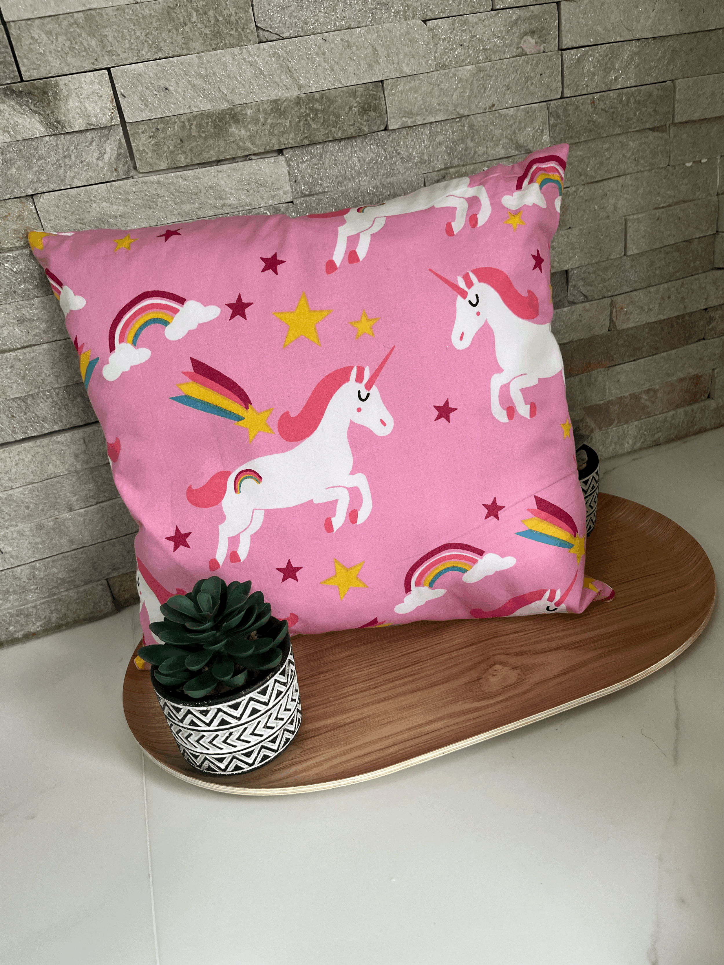 Pink cushion with unicorns, rainbows, stars, and clouds design, placed on a wooden tray with a small potted succulent plant, against a stone wall background.
