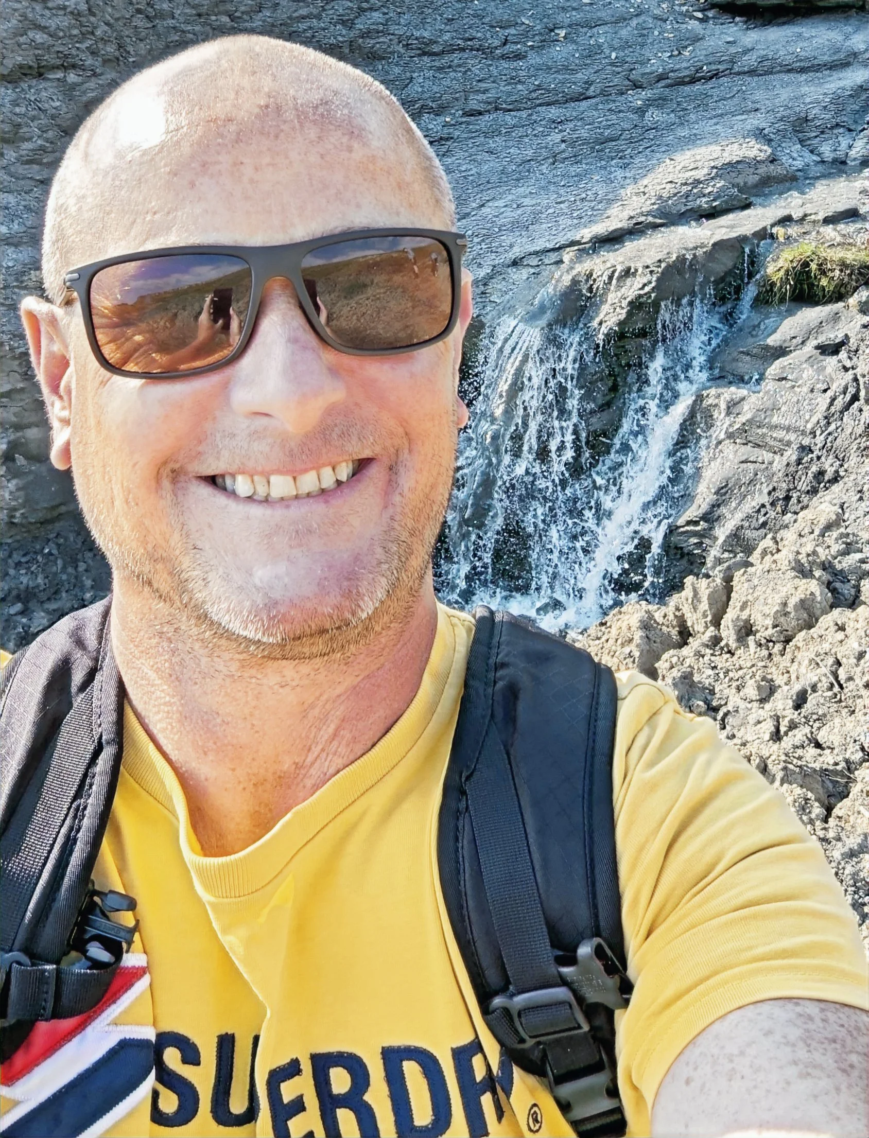 Person taking a selfie in a rocky outdoor setting with a small waterfall in the background, wearing sunglasses, a yellow shirt, and a backpack.