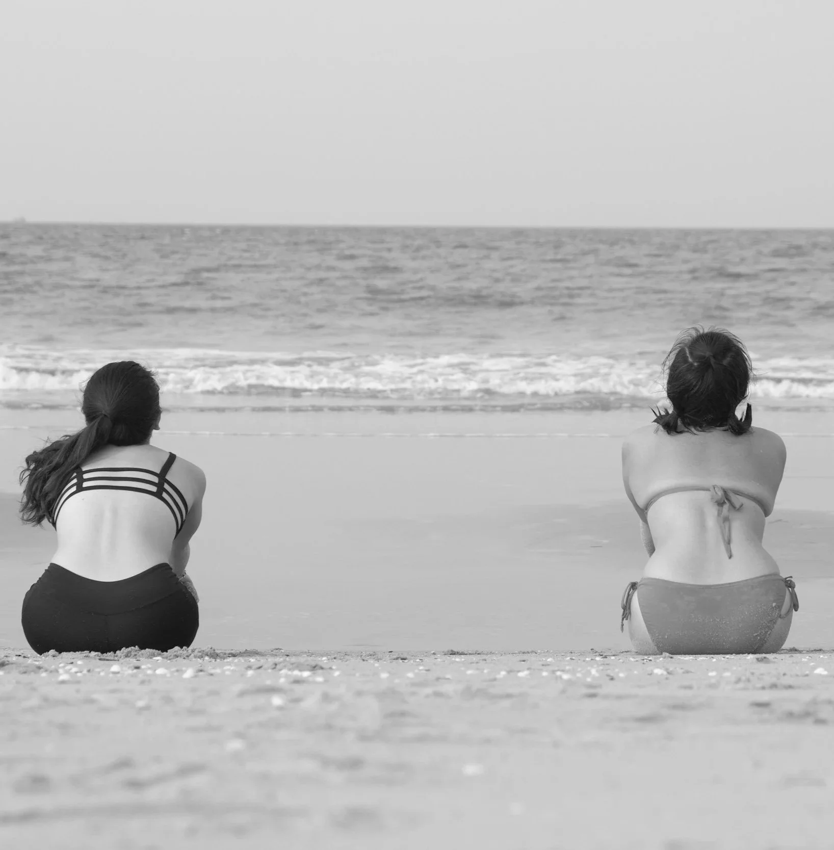 Two girls sitting on a beach facing the ocean, one with long hair and the other with two pigtails, viewed from behind.