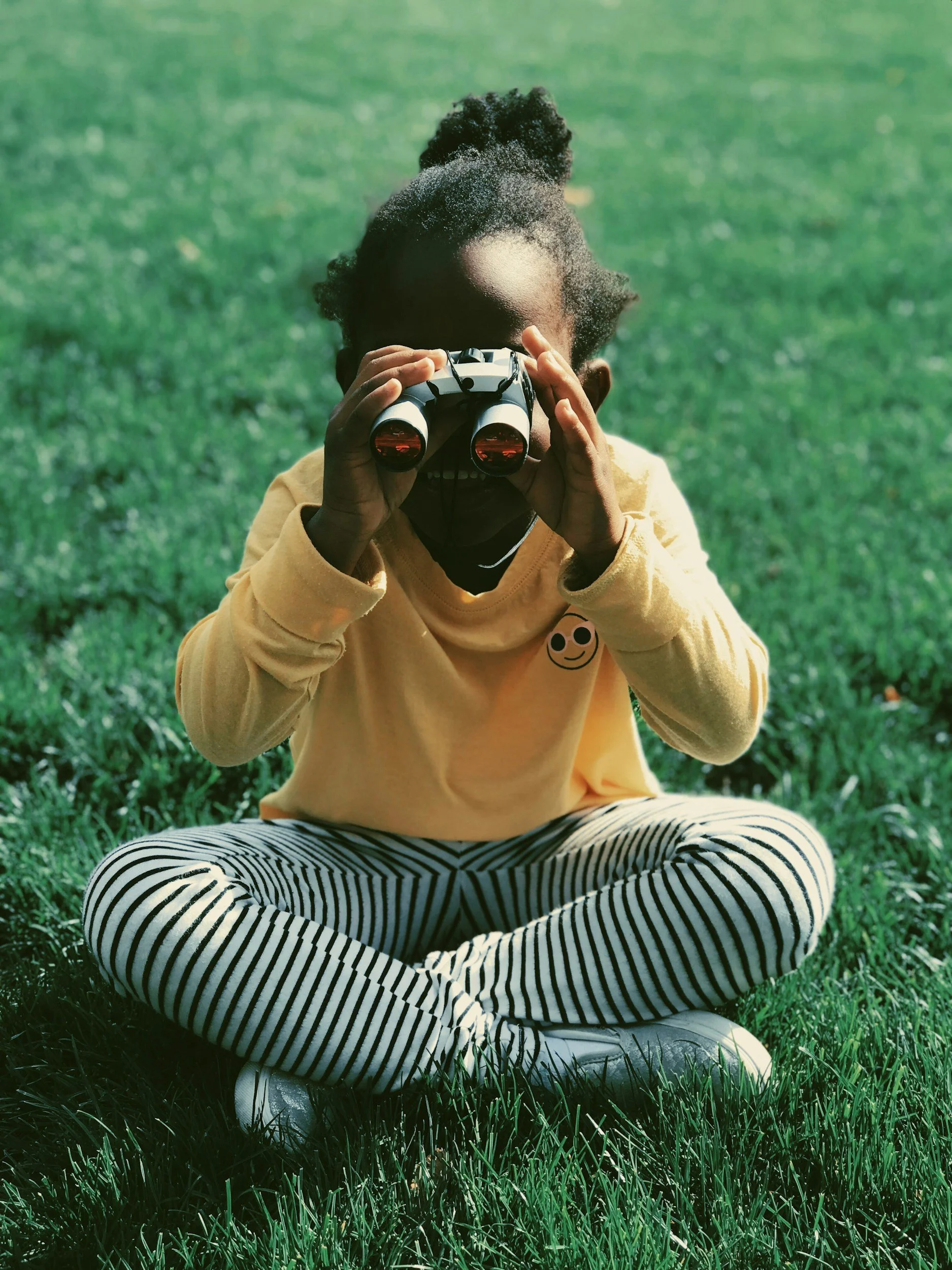 Child sitting cross-legged on grass, holding binoculars and looking through them, wearing a yellow sweatshirt with a smiley face, striped pants, and white sneakers.