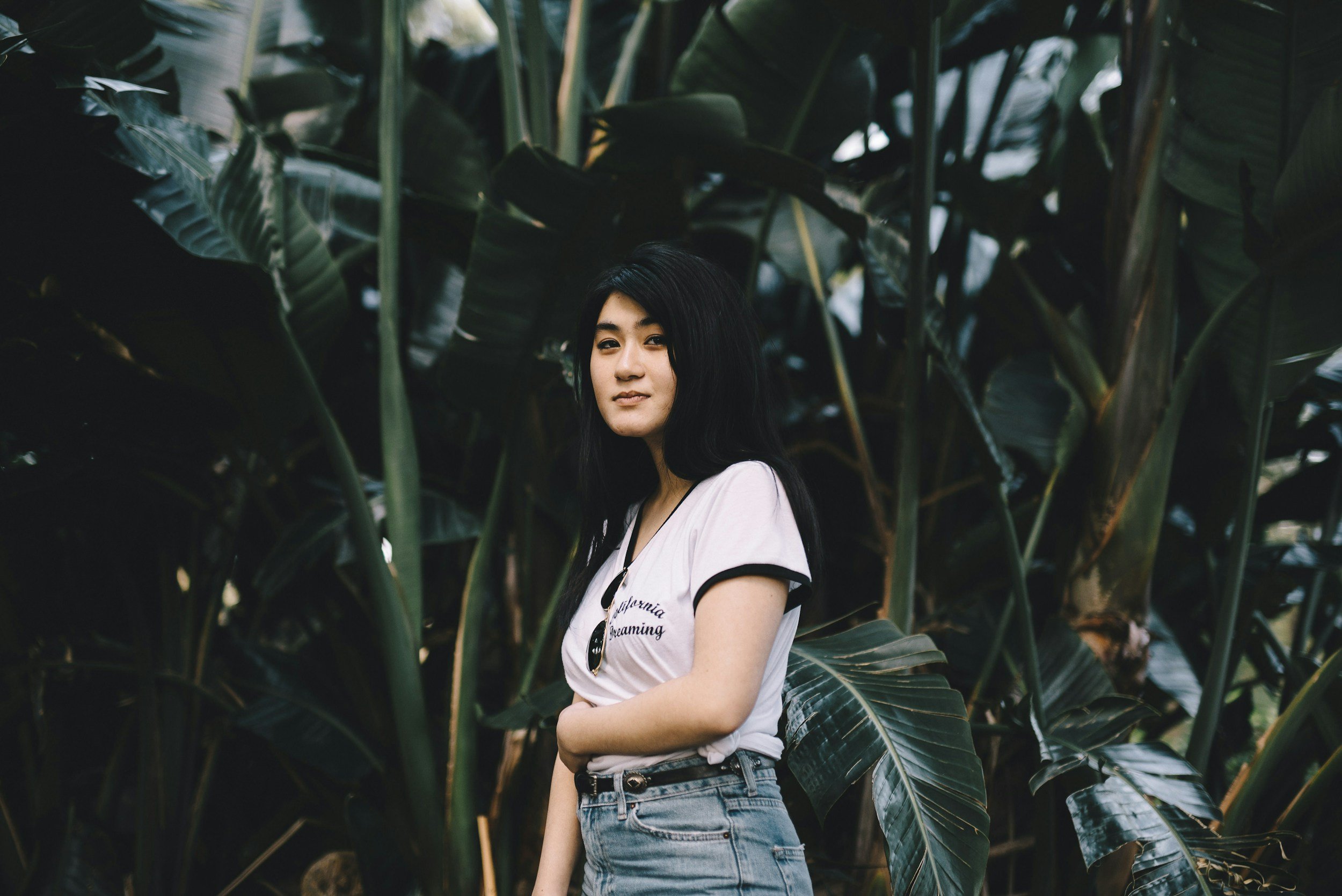 A young woman with long black hair standing among tall green tropical plants, wearing a white t-shirt and denim shorts, looking at the camera with a neutral expression.