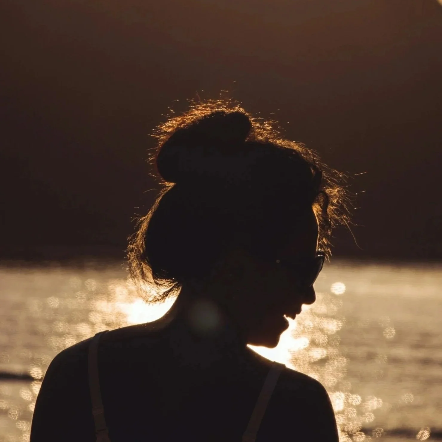Silhouette of a woman with sunlight reflecting on water behind her, with her hair tied up, at the beach during sunset.