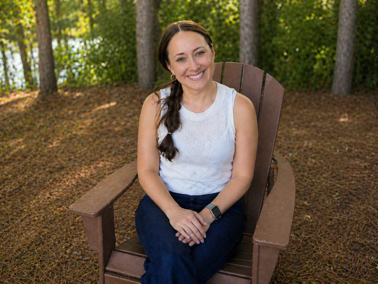 A woman with long brown hair in a braid, wearing a white sleeveless top and dark jeans, sits on a wooden Adirondack chair in a wooded area with a forest background. She is smiling at the camera.