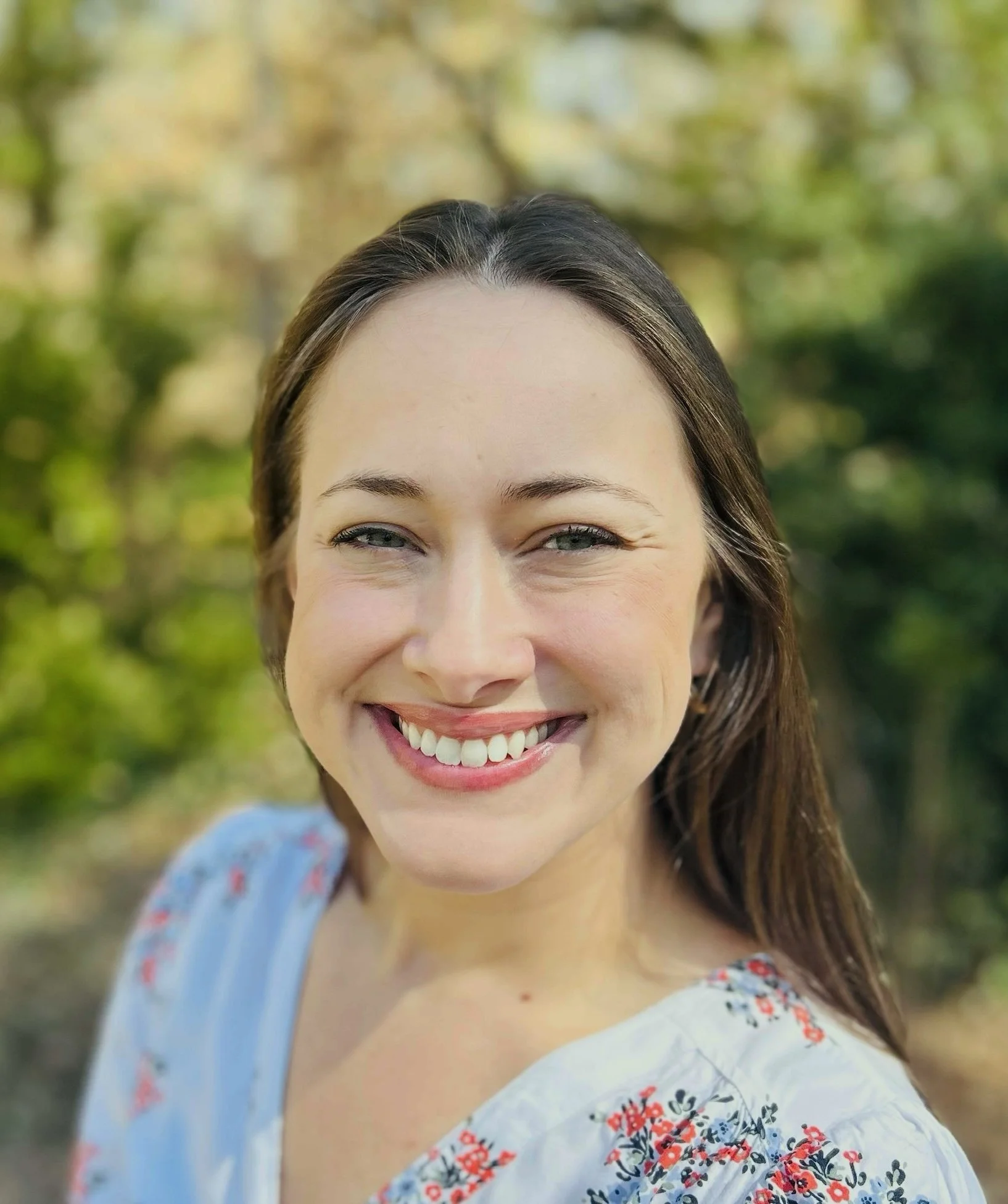Close-up of a smiling woman with brown hair and blue eyes outdoors with blurred green foliage in the background.