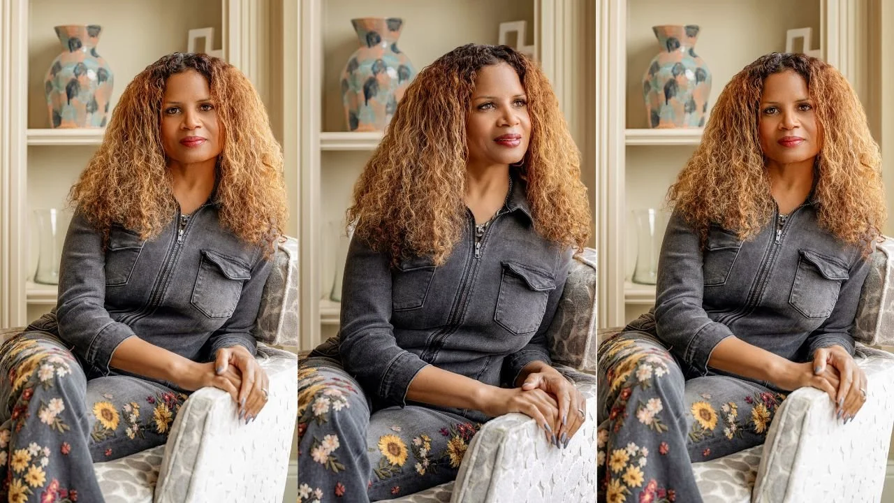 A business woman with curly reddish-brown hair sitting in armchair getting her portrait taken in front of a beige wall with shelves and decorative vases with natural light.