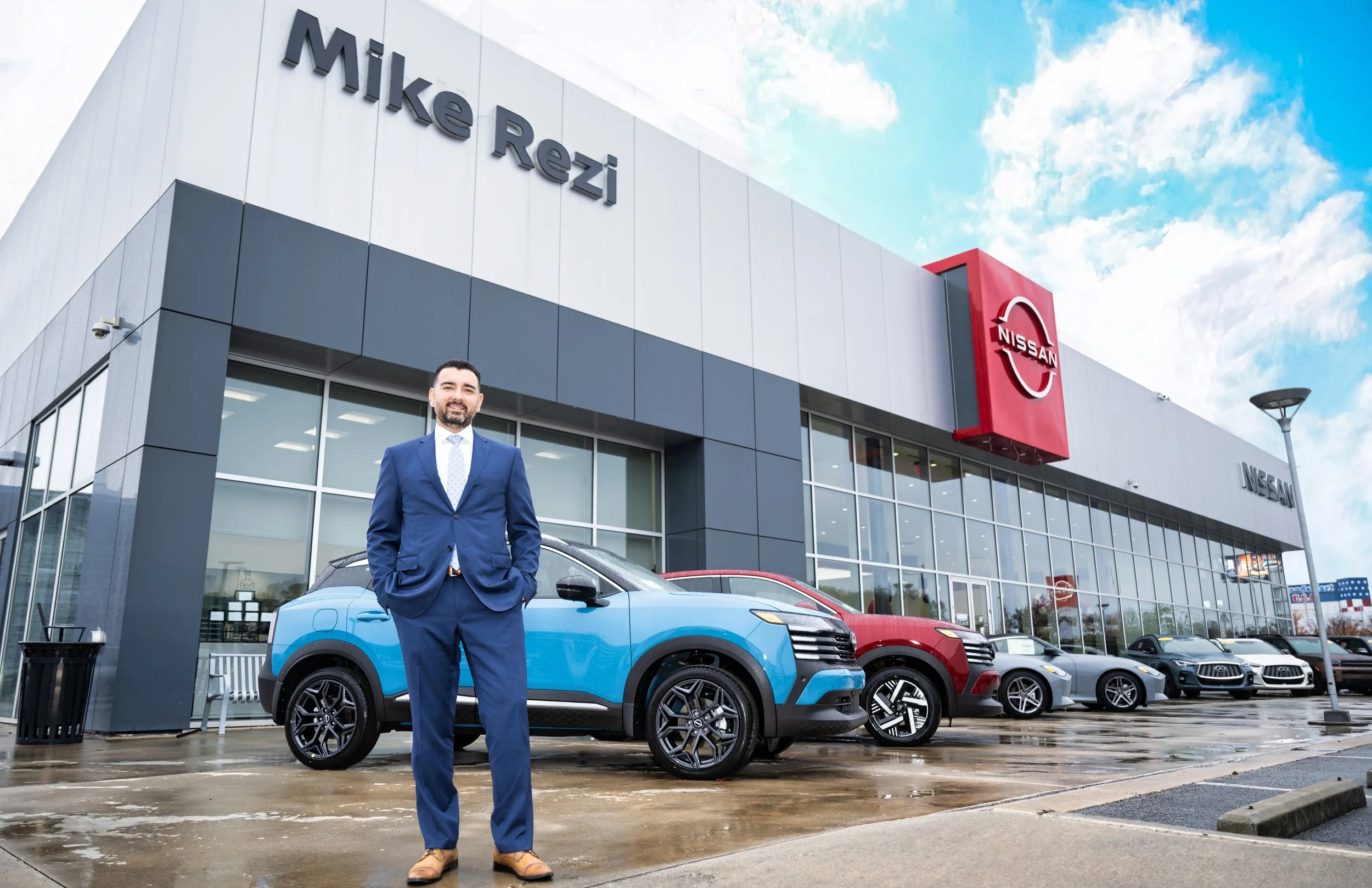 Person in a blue suit standing outside a Nissan dealership, with a few cars parked in front of the large glass building.