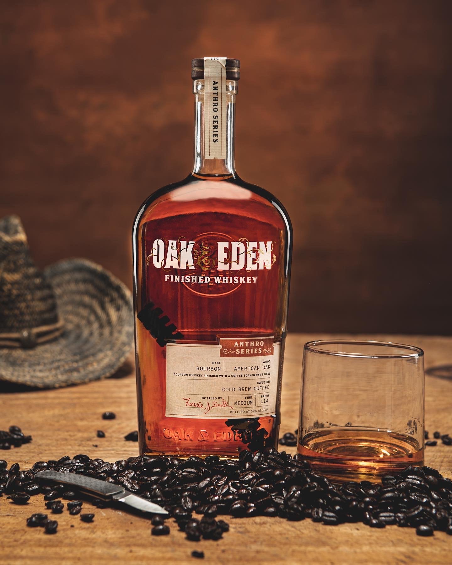 Oak & Eden whiskey bottle with glass of whiskey and coffee beans on wooden table, cowboy hat in background.