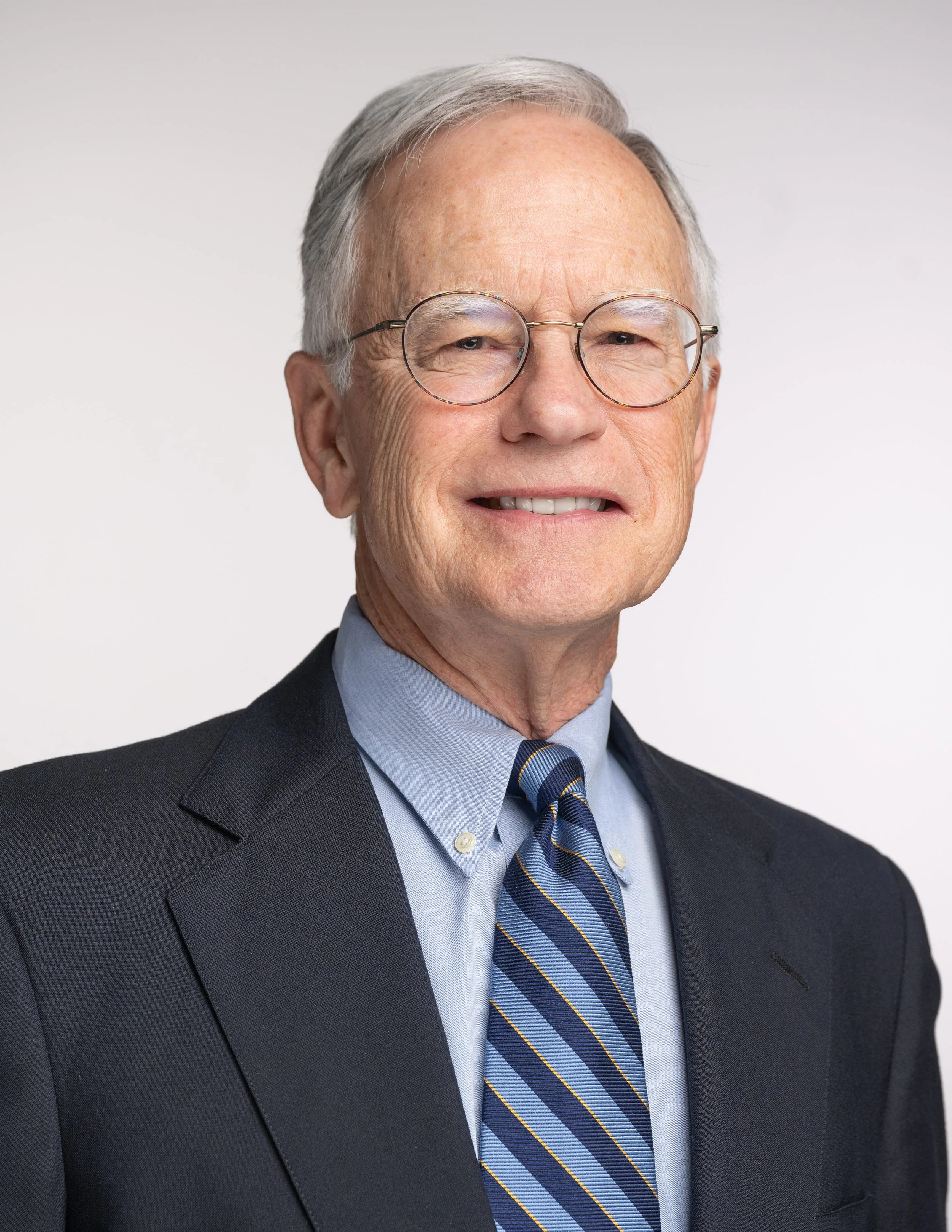 A older man with gray hair, glasses, and a suit, smiling getting his headshot in studio against a plain white background.