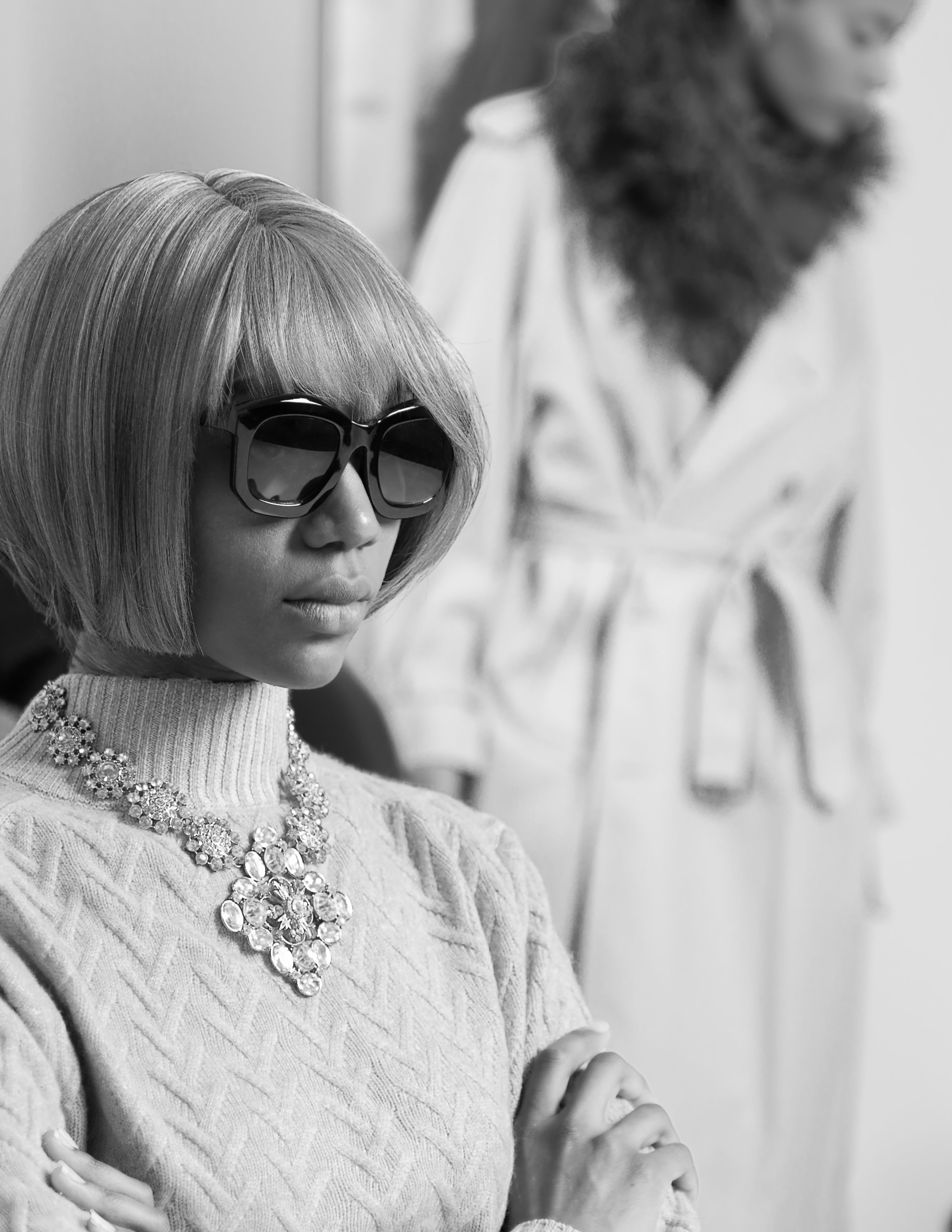 Black and white photo of a stylish woman wearing sunglasses, a bob haircut, and a statement necklace. Another woman blurred in the background, wearing a fur-collared coat.