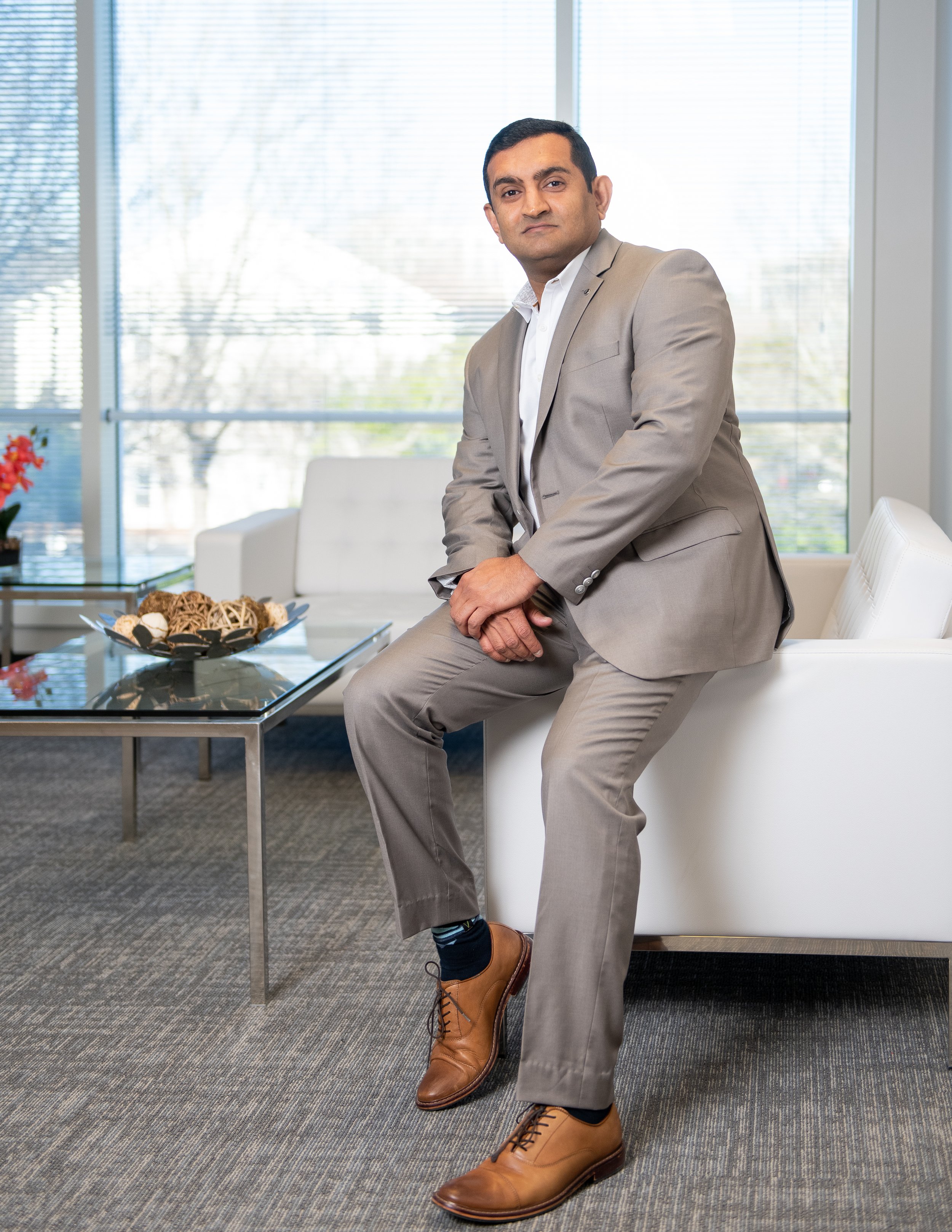 Man in a beige suit sitting on a white sofa in an office setting with a glass table and decorative items.