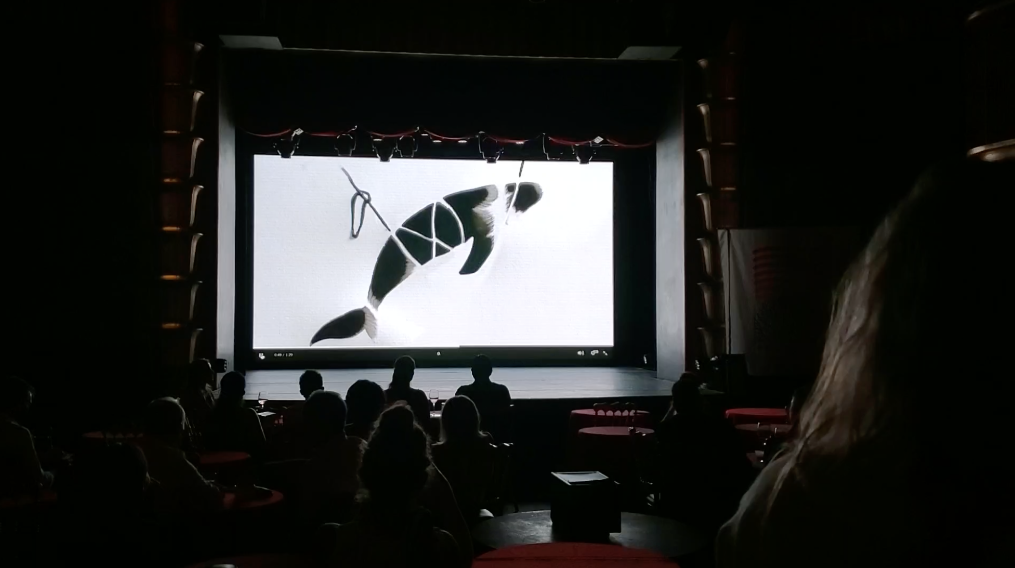 An audience watching a large screen in a theater showing a shadow silhouette of a fish harnessed in a net. The theater is dimly lit with visible silhouettes of people sitting at tables.