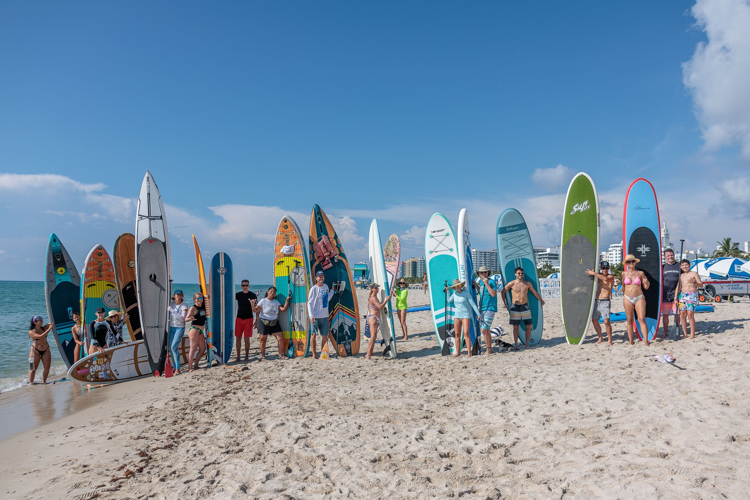 Group of people posing with stand-up paddleboards on a beach with ocean and buildings in the background.