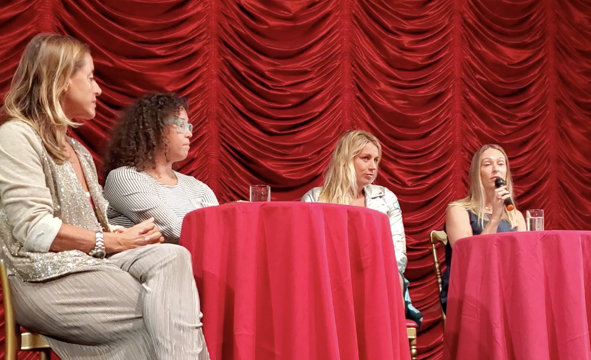 Four women seated at a round table with a red tablecloth, in front of a red curtain. One woman is speaking into a microphone.