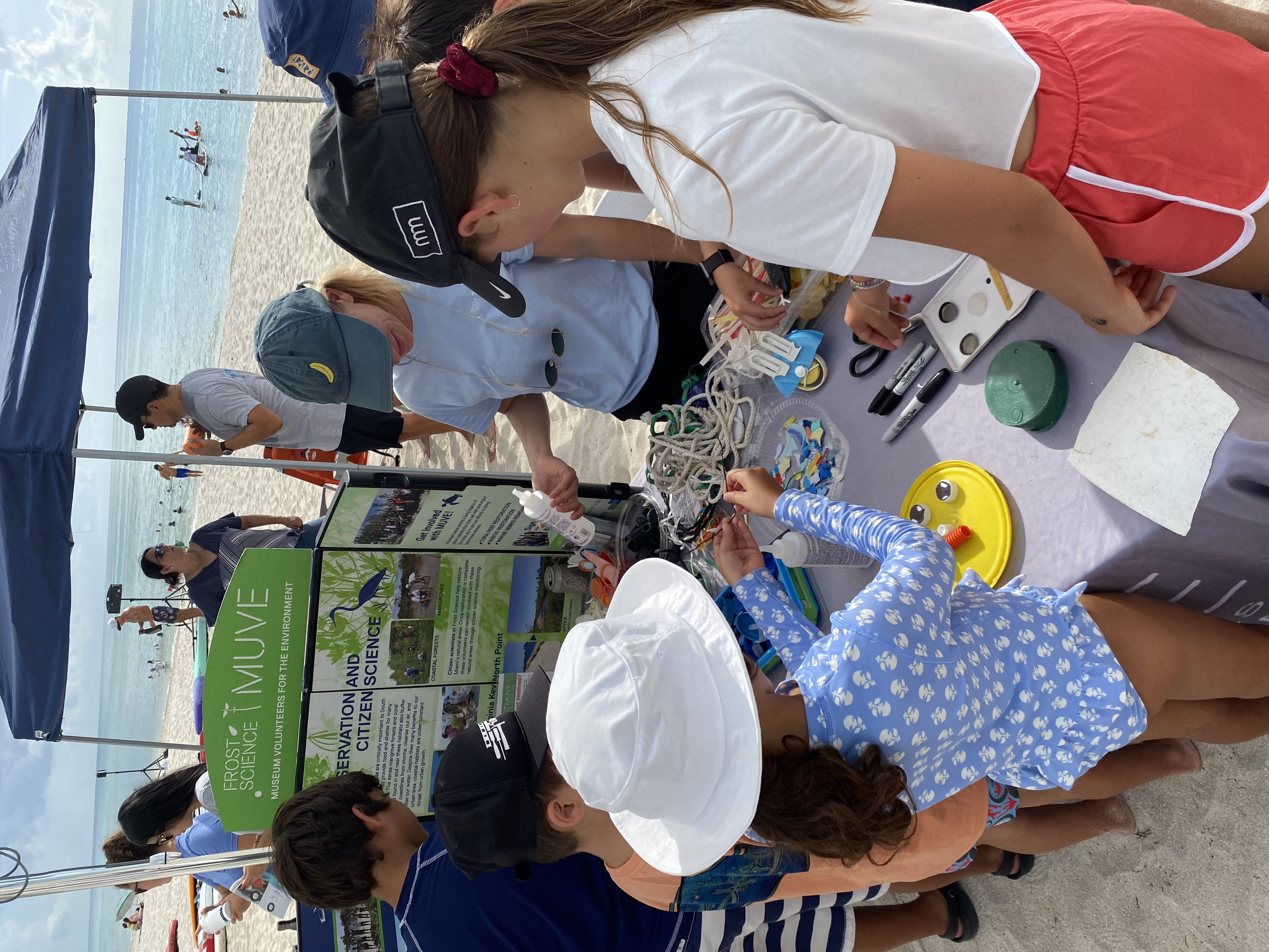 People gathered at a beach-side table under a canopy, engaging in educational activities related to environmental conservation. The table displays informational materials and crafting supplies. Paddleboarders are visible in the ocean.