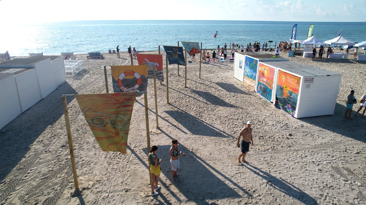 Beach art installation with colorful flags and murals, featuring people walking and standing on the sand near the ocean.