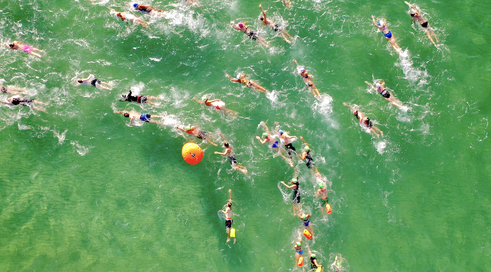 Aerial view of a group of swimmers participating in an open water swimming race in a green body of water, with an orange buoy marking the course.