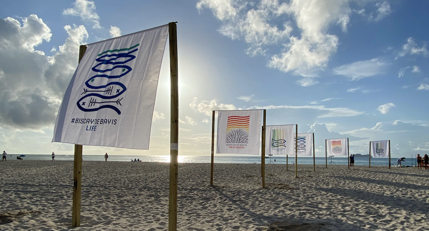 Beach with flags displaying various designs promoting Biscayne Bay, people walking, and sunlight reflecting on the ocean.