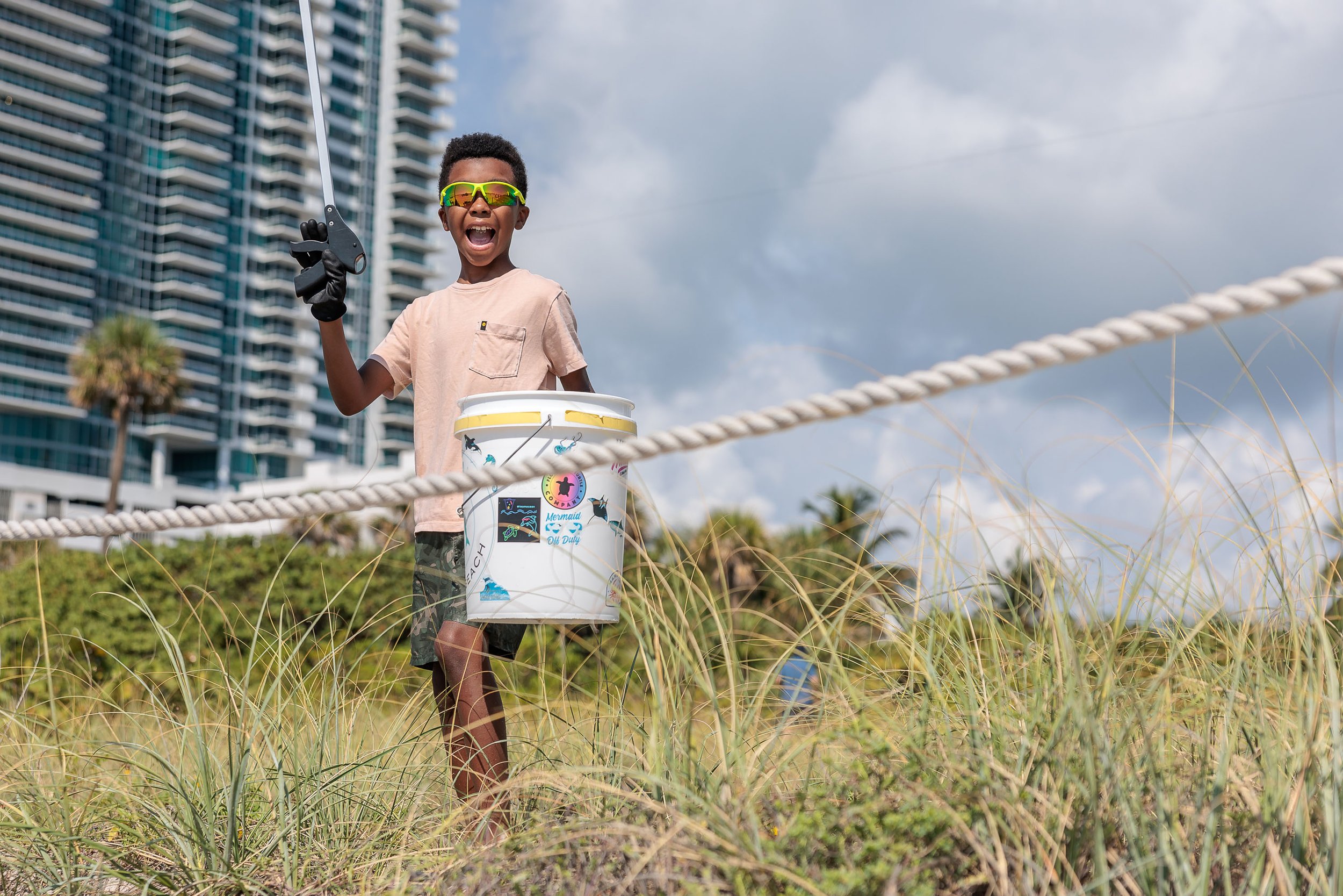 Child in sunglasses holding a trash picker and bucket, cleaning beach with buildings in background.