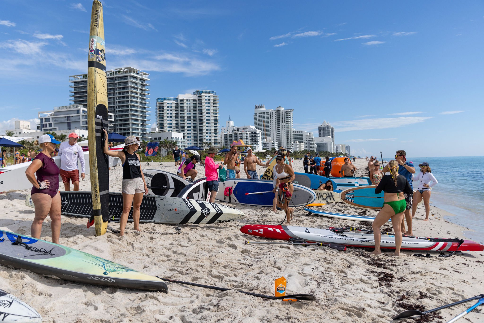 People gathered on a beach with paddleboards, skyscrapers in the background.
