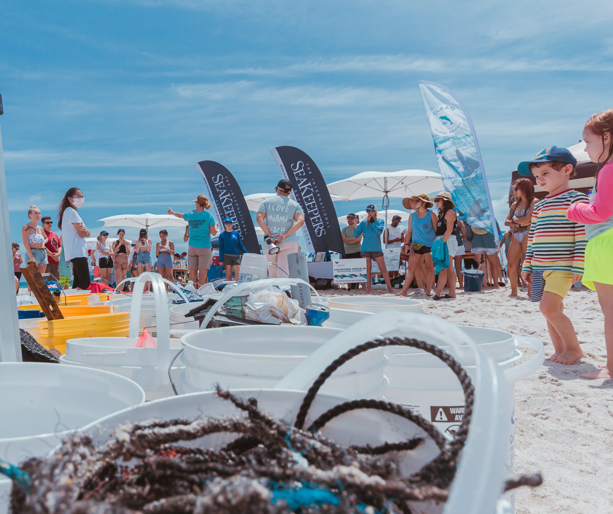 Beach cleanup event with participants gathering, buckets for trash, and banners.