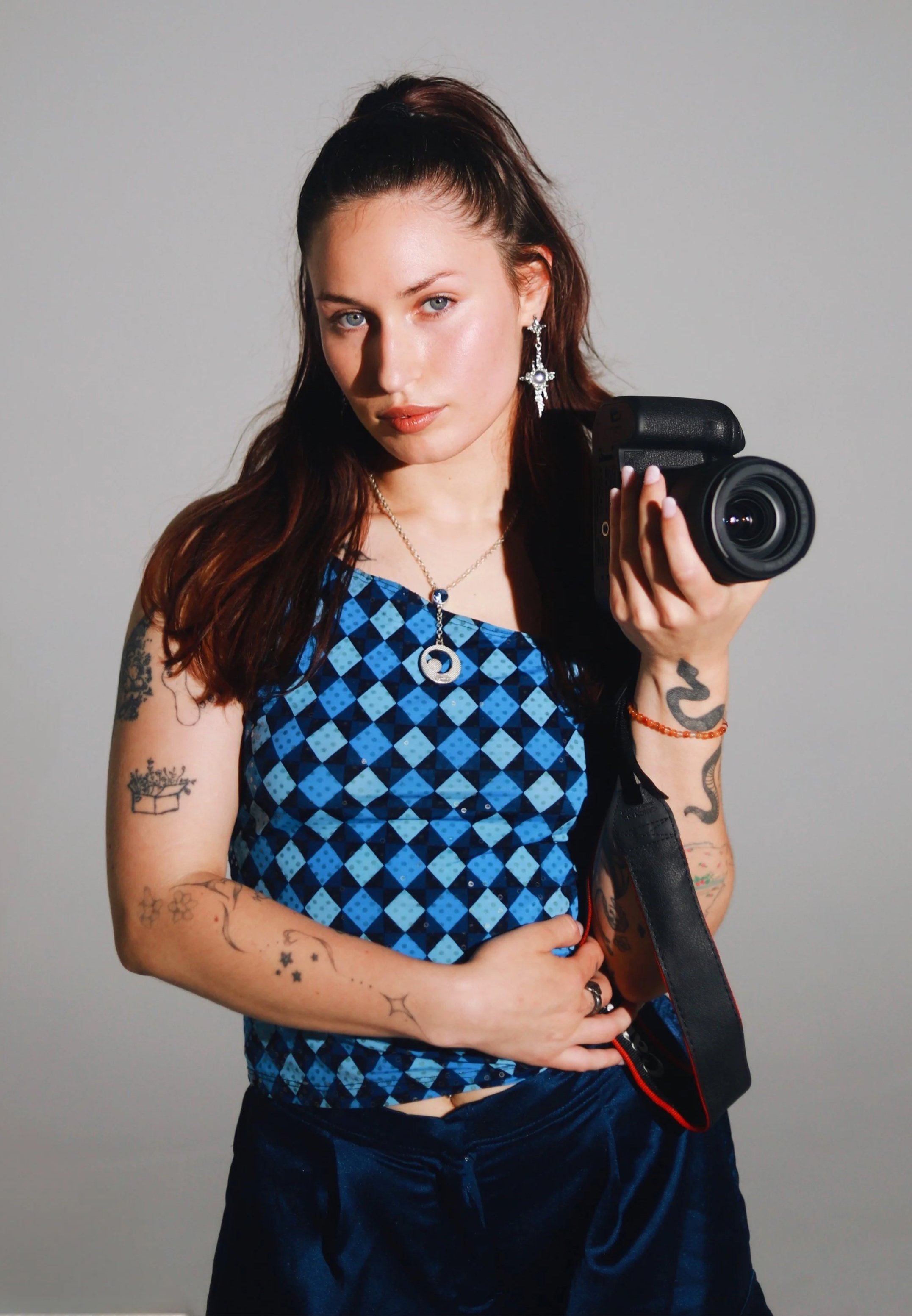 A young woman with long brown hair, tattoos on her arms, wearing a blue patterned sleeveless top, is taking a selfie with a professional camera against a plain background.