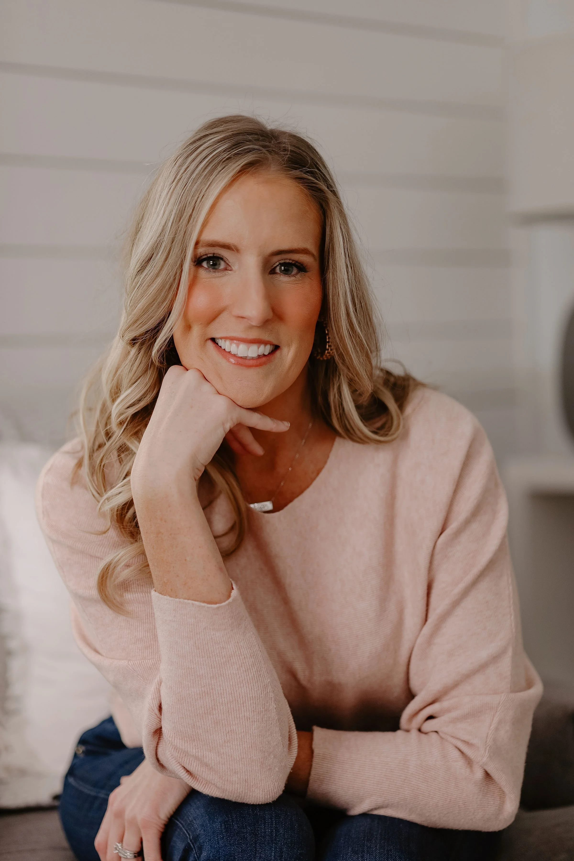 A woman with blonde hair and a bright smile, sitting indoors with a white wall background, wearing a light pink sweater and jewelry.