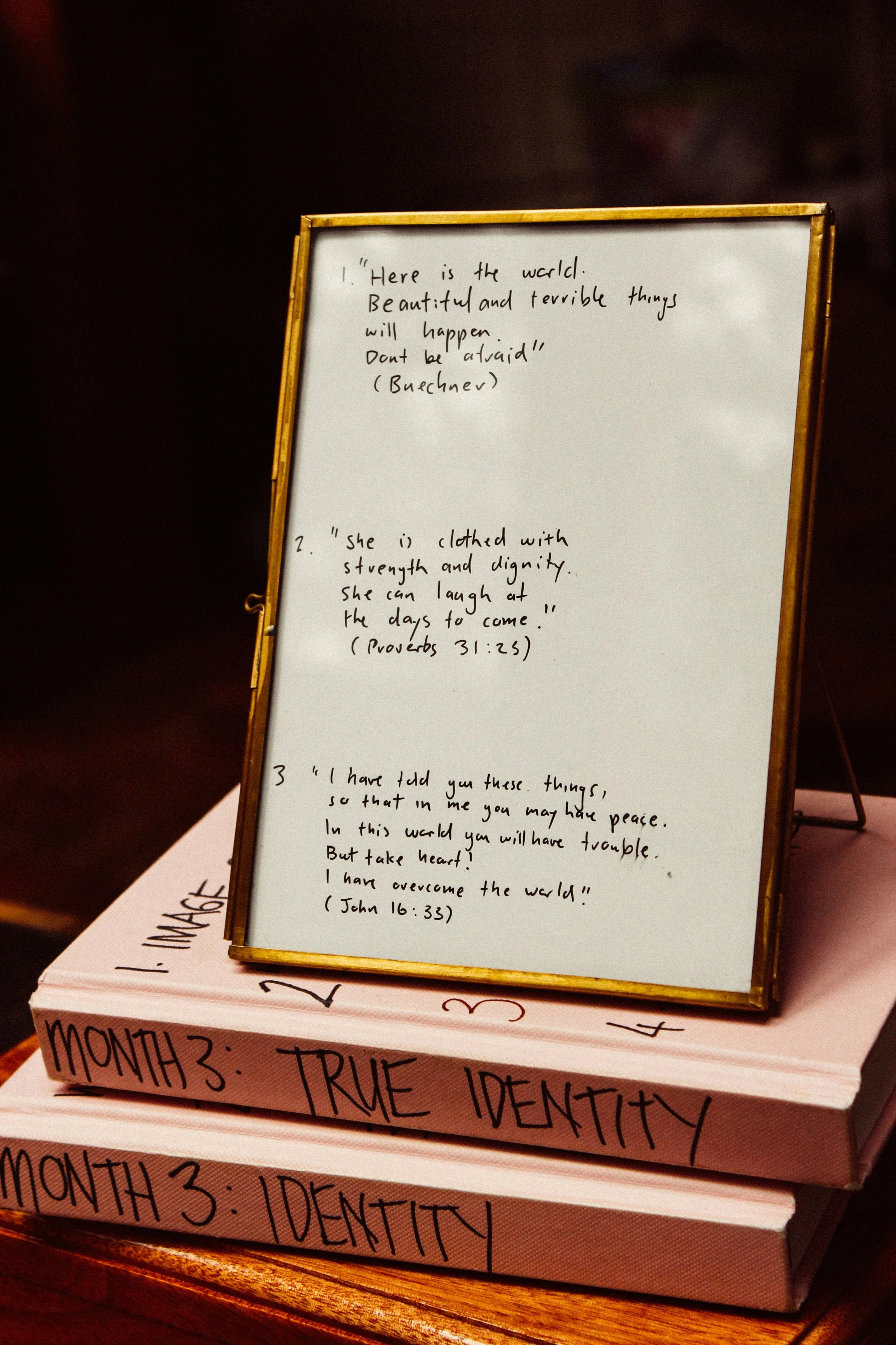 A framed paper with three quotes written in black ink; it rests on books labeled 'Month 3: True Identity' and 'Month 3: Identity.'