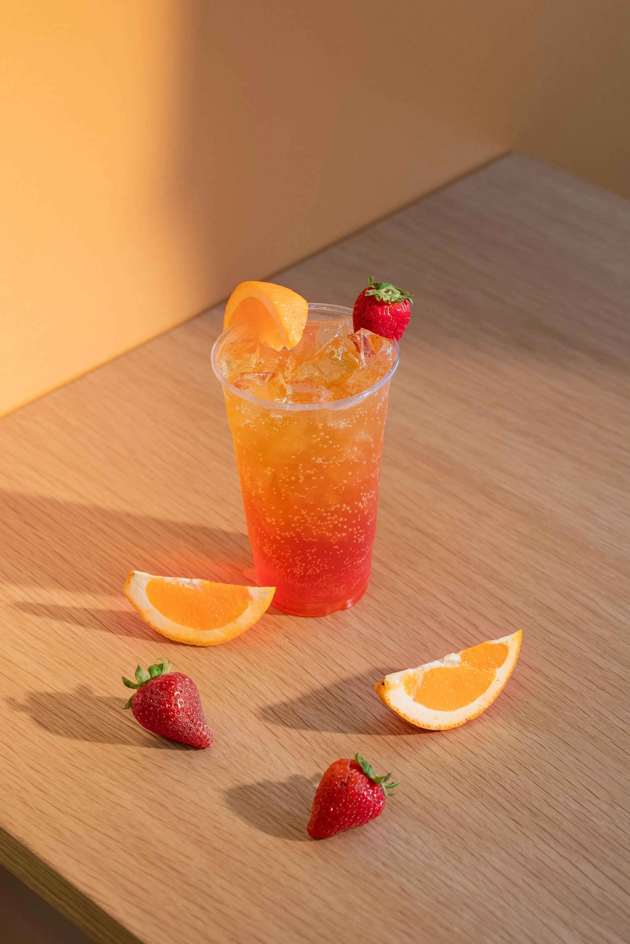 A tall glass of strawberry-orange soda with ice, garnished with a strawberry and an orange wedge on top, placed on a wooden surface with additional strawberries and orange wedges around it.